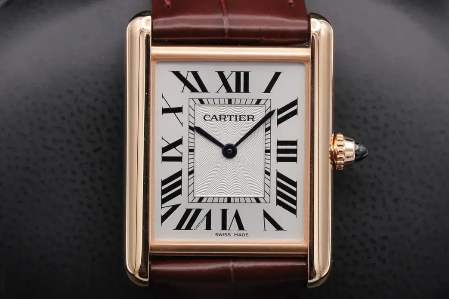 A close-up of a Cartier Tank watch with a rectangular gold case, white dial, black Roman numerals, blue hands, and a brown leather strap. The crown has a blue gemstone. The dial reads Cartier and Swiss Made.