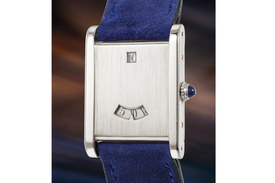 A close-up of a rectangular wristwatch with a brushed metal face, digital hour and minute windows, and a blue suede strap. The watch crown features a blue cabochon. The background is softly blurred.