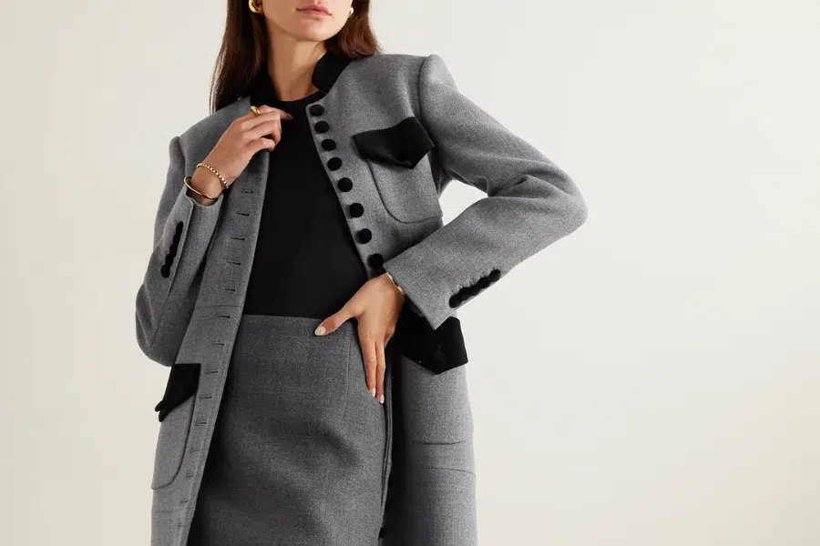 Woman wearing a tailored gray blazer with black buttons and accents, paired with a matching gray skirt and black top, posing with one hand on her hip against a plain background.