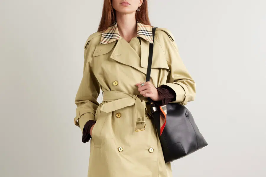 A woman wearing a beige trench coat with a plaid collar detail stands against a plain background, carrying a black shoulder bag with a colorful scarf tied to it.
