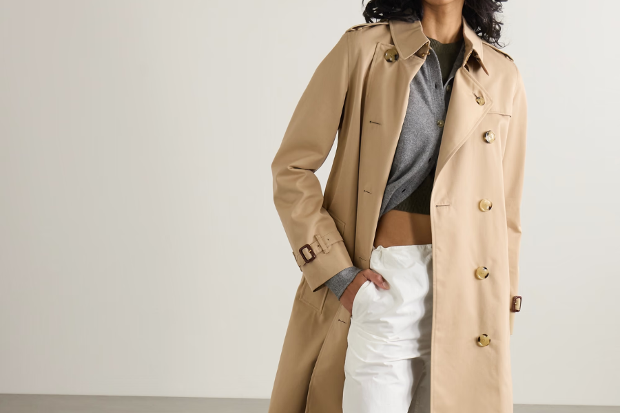 A person wearing a beige trench coat over a gray cropped shirt and white pants stands with one hand in their pocket against a plain light background.