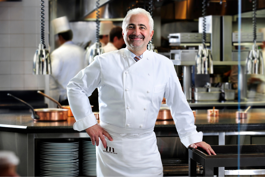 A smiling male chef in a white uniform stands confidently in a professional kitchen with other chefs working in the background, surrounded by stainless steel equipment and copper pots.