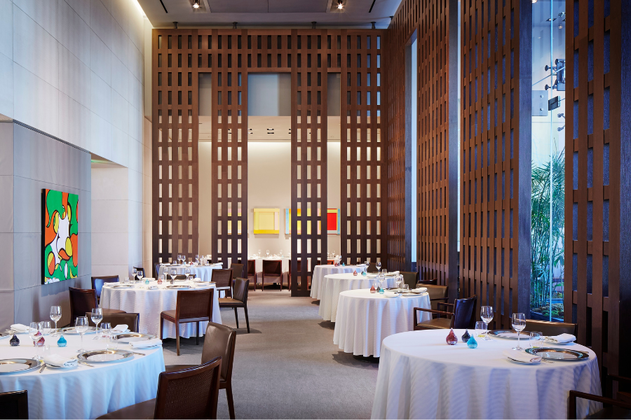 A modern, elegant restaurant with round tables covered in white cloths, brown chairs, abstract art on the walls, and tall wooden partitions dividing the space. The atmosphere is bright and sophisticated.