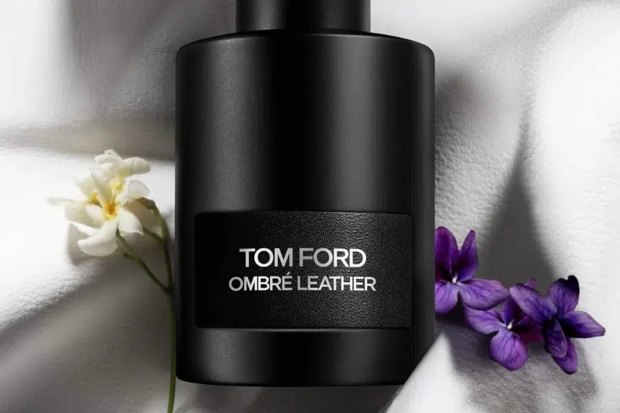 A black Tom Ford Ombré Leather perfume bottle is set on a white fabric background, with a small white flower and purple flowers beside it.