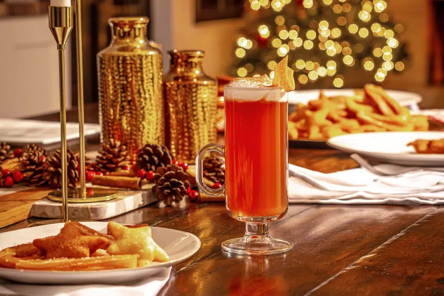 A festive table setting with a glass of red drink topped with foam, plates of star-shaped cookies, pinecones, candles, and golden decorations. A lit Christmas tree glows softly in the background.
