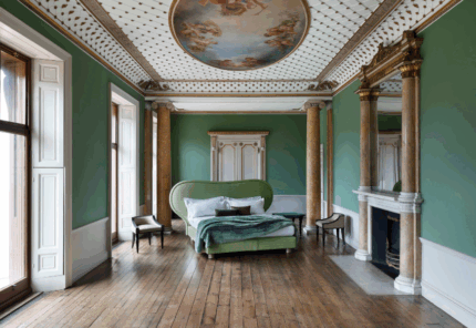 A grand bedroom with green walls, a large ornate bed, wooden floor, marble fireplace, gilded mirror, tall windows, and a painted ceiling featuring classical figures. The room has elegant, classical decor.