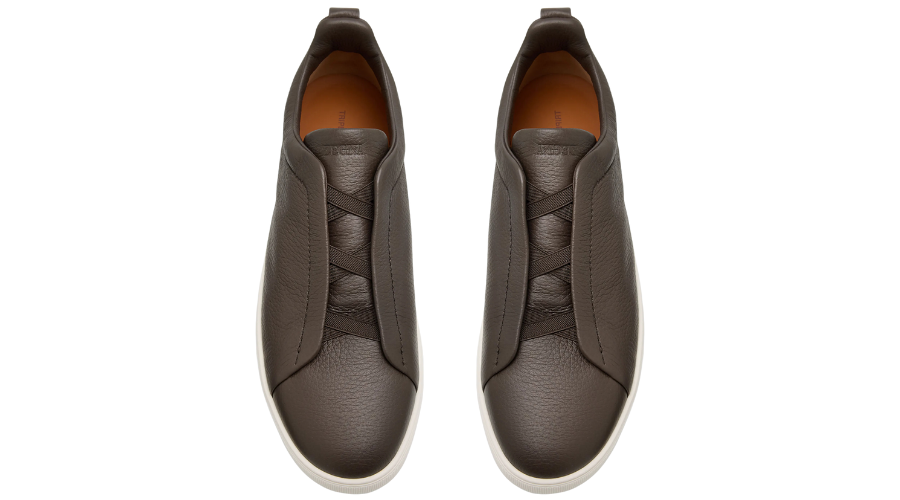 A top-down view of a pair of brown leather slip-on sneakers with white soles and elastic crossing bands over the instep. The shoes are placed side by side on a white background.