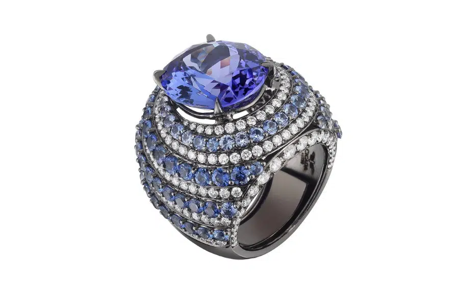 A luxurious ring featuring a large, cushion-cut blue gemstone at the center, surrounded by rows of smaller blue and clear stones set in a bold, dark metal band.