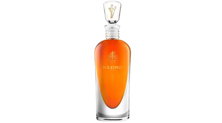 A clear glass bottle filled with amber liquid, labeled Máximo with a gold logo on the front. The bottle has a rounded, elegant shape and a transparent stopper with a gold accent inside.