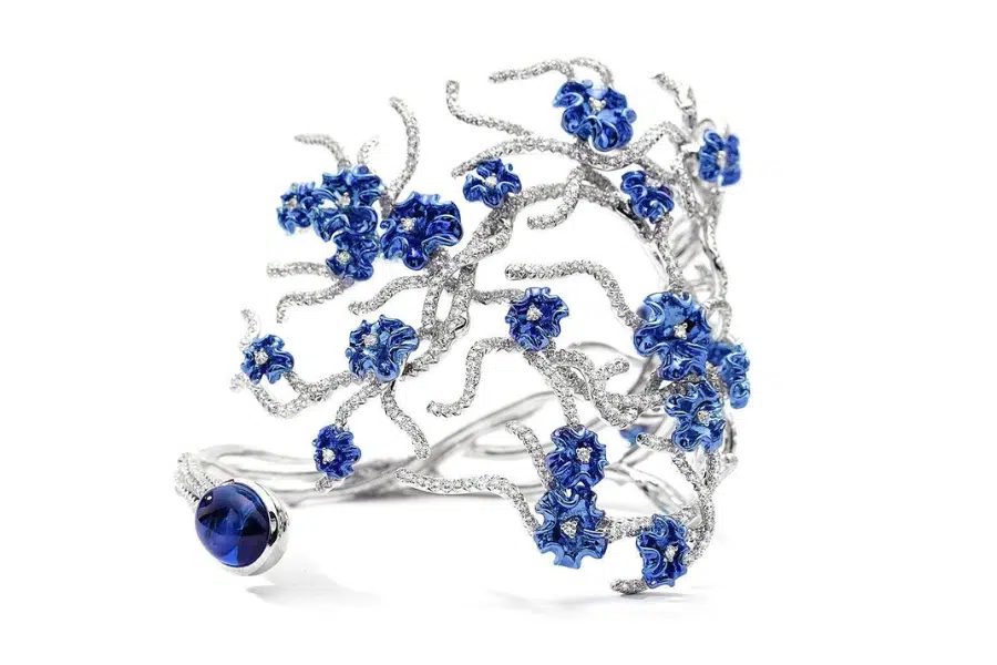An intricate silver bracelet designed like twisting branches, adorned with blue flower-shaped gemstones and a large round blue gem on one end, set against a white background.