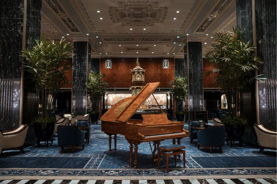 A luxurious hotel lobby with a grand piano at the center, surrounded by elegant armchairs, tall marble columns, large potted plants, and an ornate clock in the background beneath an intricate ceiling.