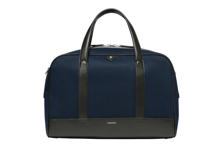 A navy blue weekender bag with black leather handles, black leather accents, and a small metallic logo at the top center. The bag is structured and has a zippered closure.