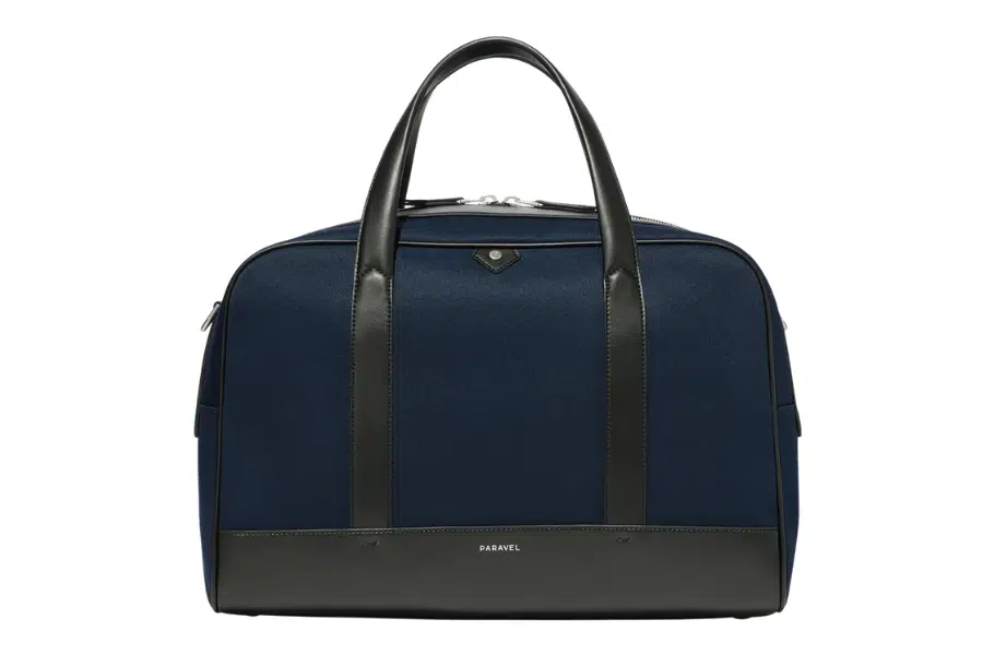 A navy blue weekender bag with black leather handles, black leather accents, and a small metallic logo at the top center. The bag is structured and has a zippered closure.