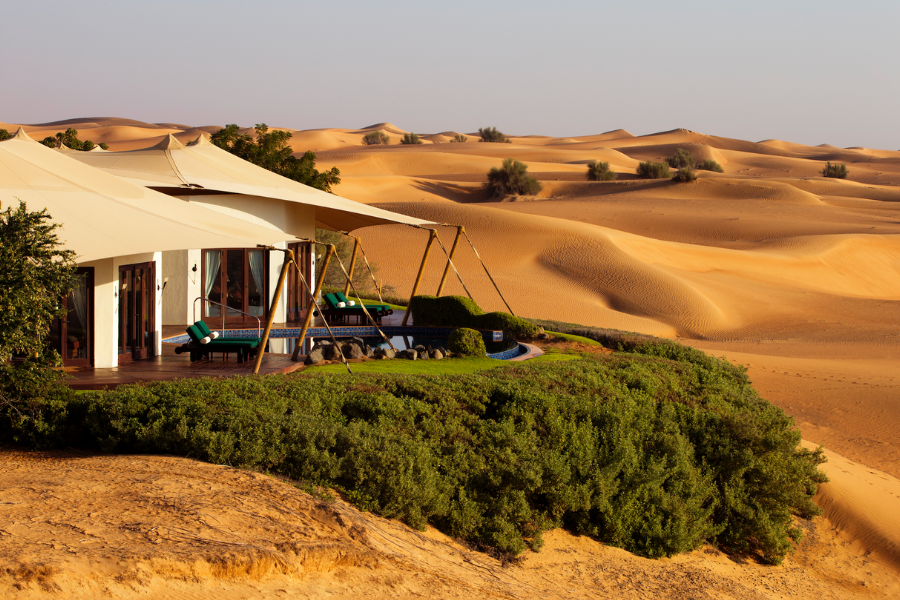 A luxury tented villa with a shaded deck, lounge chairs, and a swing sits on a lush green hill, overlooking vast golden sand dunes in a desert landscape under a clear sky.