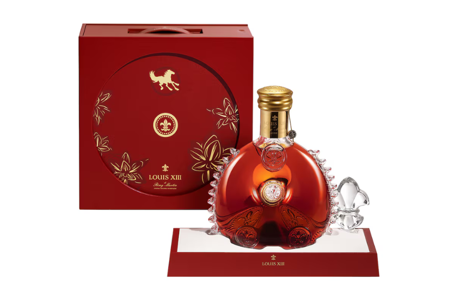 A bottle of Louis XIII cognac with a gold cap sits on a red and white display tray, next to an ornate red box decorated with gold floral designs and a horse emblem.
