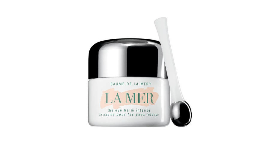 A jar of La Mer The Eye Balm Intense with a shiny silver lid, alongside a white-handled applicator with a rounded metal tip, displayed on a white background.
