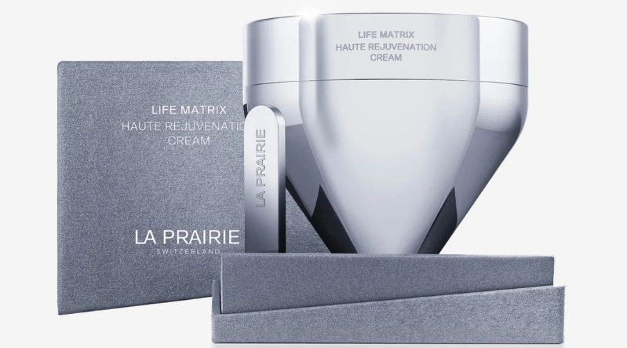 A sleek, silver jar labeled Life Matrix Haute Rejuvenation Cream by La Prairie stands next to its gray textured box, with a matching spatula resting alongside the packaging.