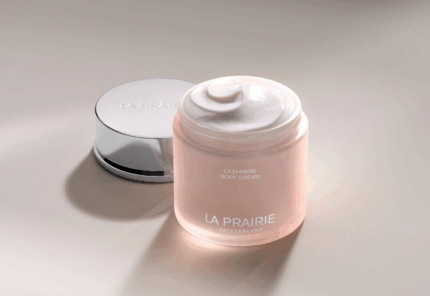 A jar of La Prairie Cashmere Body Cream with its shiny silver lid placed beside it, displaying a swirl of creamy white product inside, set on a smooth, light-colored surface.