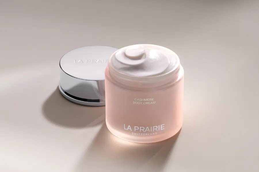 A jar of La Prairie Cashmere Body Cream with its shiny silver lid placed beside it, displaying a swirl of creamy white product inside, set on a smooth, light-colored surface.