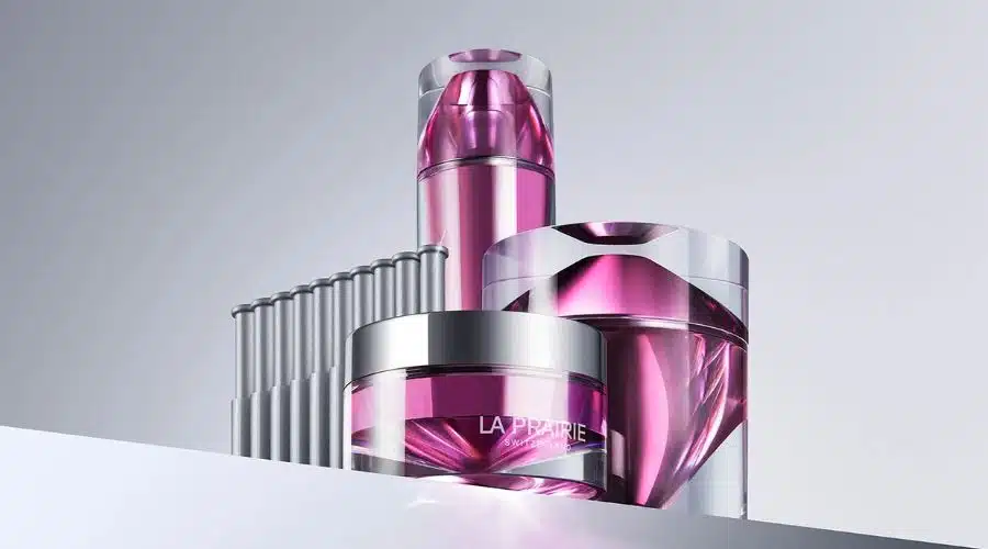 Three elegant containers of La Prairie skincare products with purple liquid are displayed on a reflective surface against a light gray background, alongside a row of metallic tubes.