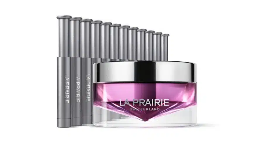 A jar of La Prairie Switzerland face cream with a silver lid and pink product inside is displayed in front of eight silver cylindrical vials, all on a white background.