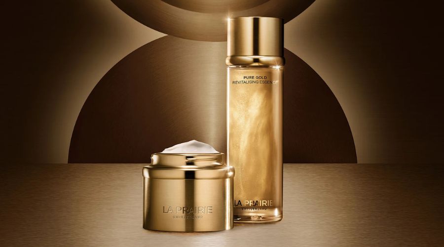 Two gold-colored skincare containers from La Prairie—one jar with a lid and cream inside, and one tall bottle—sit on a reflective brown and gold surface with an abstract circular background.