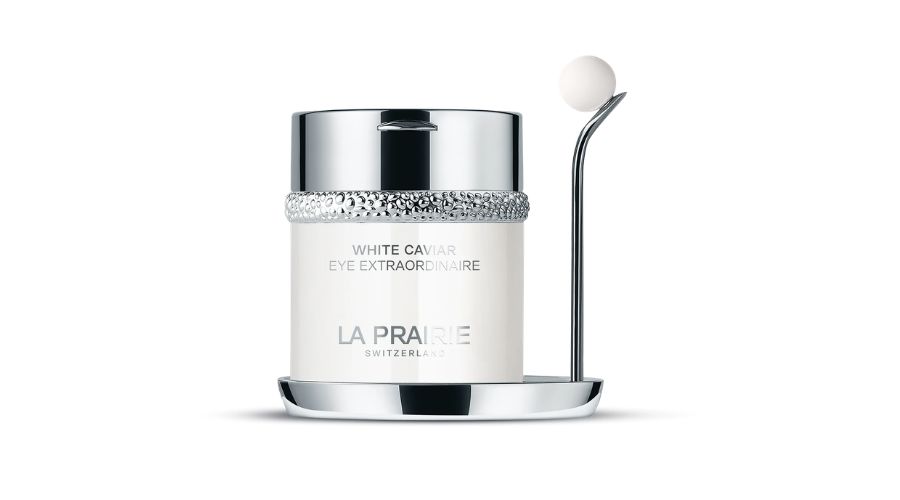 A white and silver jar labeled La Prairie White Caviar Eye Extraordinaire next to a sleek silver applicator with a white spherical tip, both placed on a matching circular tray.