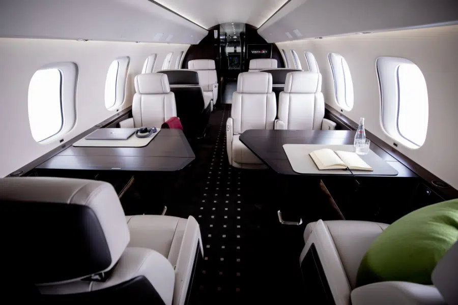 The interior of a private jet with cream leather seats, dark wood tables, a laptop, notebook, and water bottle on the tables. The cabin is well-lit with large windows on both sides.