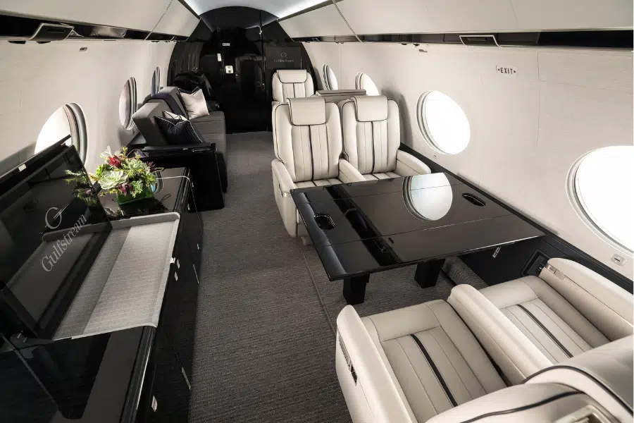 Luxurious private jet interior with cream leather seats, glossy black tables, large round windows, a sofa in the back, and a flower arrangement on a side table. The space appears spacious and elegantly designed.