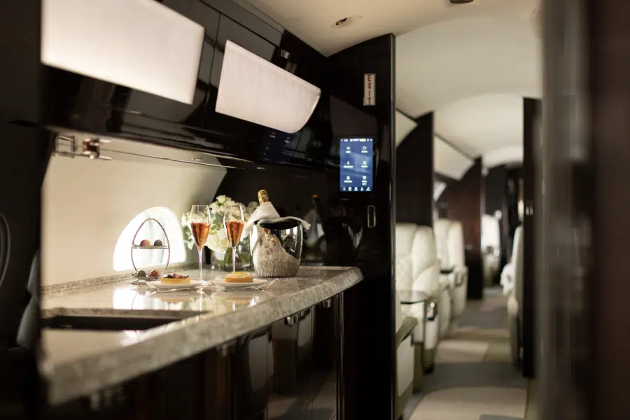 Luxurious airplane interior with a marble countertop set with champagne, glasses, and snacks. White leather seats and elegant modern decor are visible further inside the cabin.