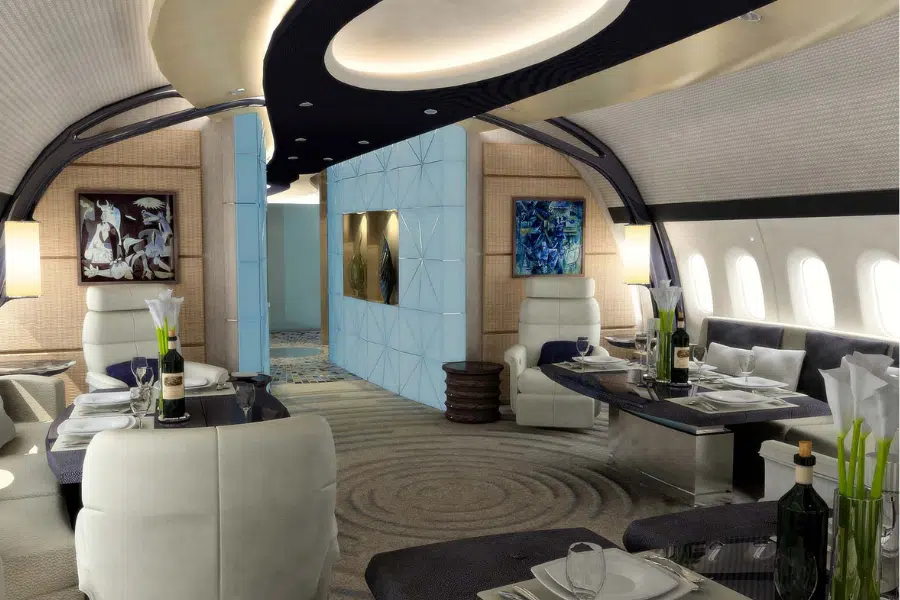 Luxurious private jet interior with plush white seating, dining tables set with wine bottles and flowers, modern lighting, abstract wall art, and large windows along one side.